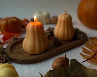 May include: Two pumpkin-shaped candles, one lit, sit on a wooden tray with pine cones. The candles are a warm orange colour, with a soft glow from the flame. Autumn leaves and pumpkins surround the tray, creating a fall theme.