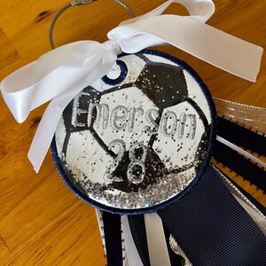 May include: A soccer-themed keychain with a white ribbon bow. The round charm features a soccer ball design with the name "Emerson" and the number "28" in silver glitter. Navy blue and white ribbons hang below.
