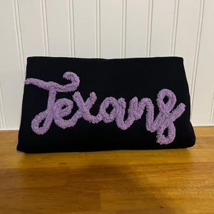 Puede incluir: Una almohada negra con la palabra "Texans" bordada en hilo de lana esponjoso morado.
