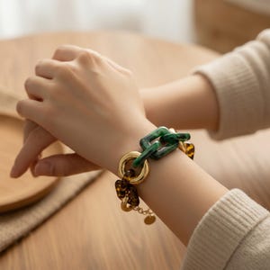 May include: A gold chain bracelet with green, tortoise shell, and gold accents. The bracelet features a large green chain link, a gold ring, and a tortoise shell link. A small gold charm hangs from the bracelet.
