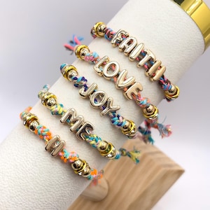 May include: Four colourful friendship bracelets are displayed. Each bracelet has a braided cord in various colours, gold beads, and gold letter beads spelling out "FAITH", "LOVE", "JOY", and "ME". The bracelets are presented on a white cylindrical stand.