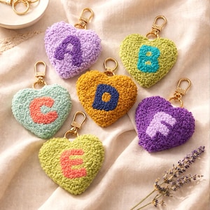 May include: Heart-shaped keychains in various colors, each featuring a different letter of the alphabet. The keychains have a textured, embroidered appearance and gold-colored hardware. The letters are in contrasting colors.