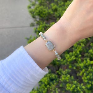 May include: A delicate bracelet with a light gray gemstone and white pearl beads. The bracelet is made with gold-toned metal.