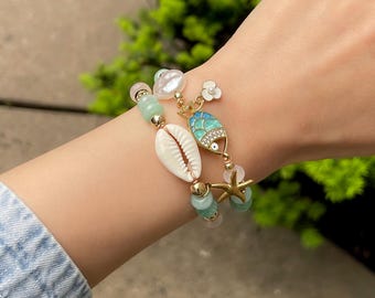 Op de afbeelding: Twee armbanden met bedels met oceaanthema. Eén armband heeft een witte zeeschelp, groene kralen en gouden accenten. De andere heeft een blauwe visbedel, een zeester en een witte bloembedel. De armbanden worden om een pols gedragen.