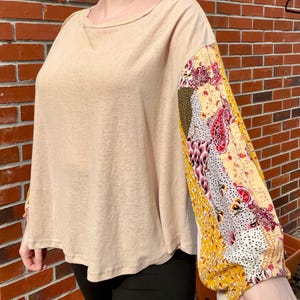May include: A beige long-sleeve top with a scoop neck and patchwork sleeves. The sleeves feature a mix of floral and paisley patterns in yellow, red, pink, and green. The top is made of a soft, lightweight material.