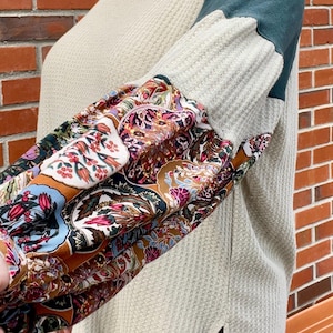 May include: A gray waffle knit top with a green raglan sleeve and a floral patterned sleeve. The top has a side slit and a relaxed fit.
