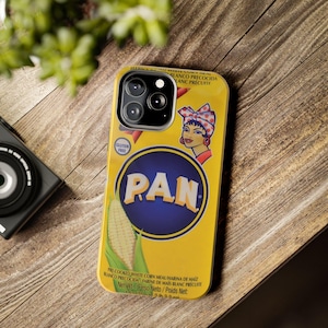 May include: Yellow phone case featuring a design inspired by a PAN cornmeal package. The case has a blue circle with the word "PAN" in white, a woman with a red headscarf, and a corn graphic. The case also has the words "GLUTEN FREE".