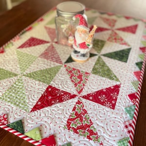 Może przedstawiać: Świąteczny bieżnik na stół z patchworkowym wzorem z czerwonych, zielonych i białych trójkątów. Bieżnik ma obramowanie w paski w stylu laski cukrowej i dekoracyjną figurkę Świętego Mikołaja. Na bieżniku znajduje się również szklany słoik.
