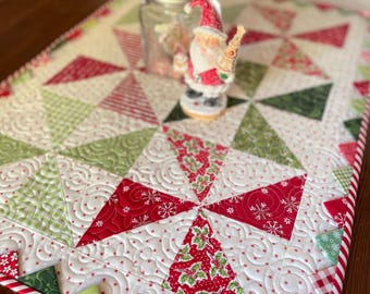 May include: A festive Christmas table runner with a patchwork design featuring red, green, and white triangles. The runner has a candy cane striped border and a decorative Santa Claus figurine. A glass jar is also on the runner.