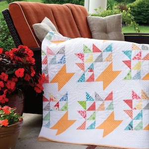May include: A white quilt with a geometric pattern of colorful triangles and orange starburst shapes. The quilt is draped over a brown outdoor sofa with pillows and red flowers in the foreground.