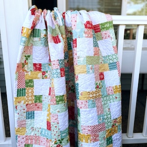 May include: A colorful patchwork quilt draped over a white railing. The quilt features a variety of patterned fabrics in red, green, yellow, and blue squares, with white quilted sections. The quilt is a mix of floral, gingham, and solid patterns.