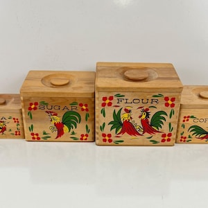 Pode incluir: Um conjunto de quatro potes de cozinha de madeira com tampas. Cada pote é pintado com um desenho de galo e rotulado com "TEA", "SUGAR", "FLOUR" e "COFFEE". Os potes são castanhos claros com detalhes em vermelho, verde e amarelo.