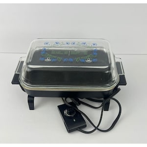 May include: Black electric skillet with a clear glass lid. The lid features a blue floral design. The skillet has a black control panel and a power cord. The appliance is designed for cooking.