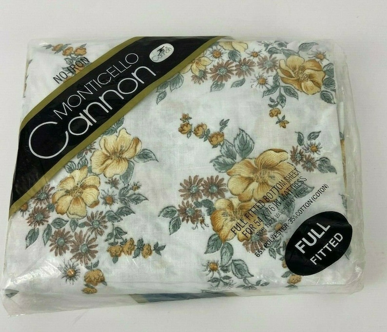 May include: A white fitted sheet with a floral pattern in shades of yellow, brown, and green. The sheet is packaged in plastic and labeled "No-Iron Monticello Cannon Full Fitted Bottom Sheet for 54 x 76 in Mattress, 65% Polyester 35% Cotton (Coton)".