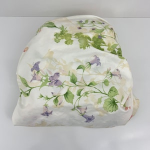 May include: White fabric with a floral pattern featuring purple bell-shaped flowers, green leaves, and light beige foliage. The fabric is folded, creating soft shadows and highlighting the delicate watercolor-style design.