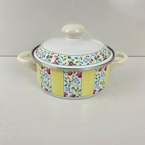 Puede incluir: Una olla de cocina de esmalte blanco y amarillo con tapa. La olla tiene un diseño floral con detalles en rojo, azul y verde. Las asas y el pomo de la tapa son de color blanquecino. La olla tiene unos 20 cm de diámetro.