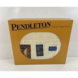 Op de afbeelding: Een crèmekleurige Pendleton-deken in een lichtbruine doos. De doos heeft het woord "PENDLETON" in donkerblauwe letters en de zin "Always Virgin Wool". De deken heeft een vierkant patroon.