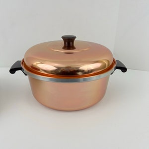 以下が含まれることがあります： 黒い取っ手とそれに合わせた蓋が付いた銅色の鍋。鍋の上部に銀色の縁取りがある。