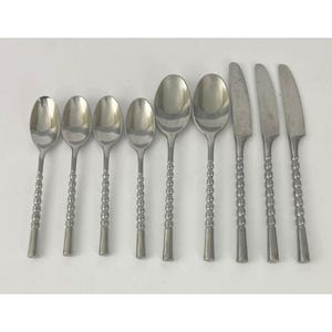 Könnte beinhalten: Ein Set aus acht Edelstahl-Besteckteilen, darunter Löffel und Messer. Die Löffel sind in verschiedenen Größen erhältlich und haben ein dekoratives Perlendesign an den Griffen. Die Messer haben ein schlichtes, elegantes Design. Das Besteck ist auf einer weißen Oberfläche angeordnet.