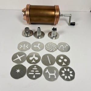 May include: A copper-colored cookie press with a silver-toned hand crank, three metal piping nozzles, and twelve circular metal discs featuring various cut-out shapes including a star, a tree, a dog, and geometric patterns.