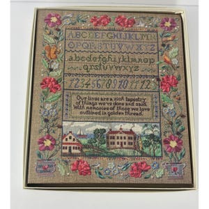 May include: A framed cross-stitch sampler with a tan background. The sampler features the alphabet, numbers, and a scenic landscape. The border is adorned with red and pink flowers, blue flowers, and green leaves. The text reads: "Our lives are a rich tapestry of things we've done and said."