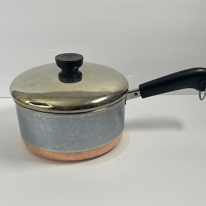 May include: A small saucepan with a copper base and a brushed silver-toned metal body. The pot features a black plastic handle with a metal hanging loop and a matching silver-toned lid topped with a round black plastic knob for easy handling.