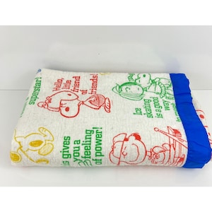 Puede incluir: Manta de vellón blanca con temática de Peanuts. La manta presenta varios personajes y citas de Peanuts, incluyendo Snoopy, Charlie Brown y Lucy. La manta tiene un borde de satén azul.