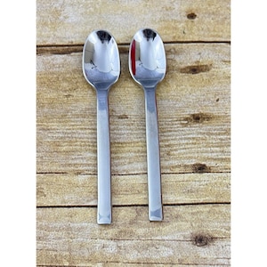 May include: Two stainless steel spoons with oval bowls and long, rectangular handles. The spoons are highly reflective, mirroring the surroundings. They rest on a light brown, wooden surface, likely a table or countertop. The spoons are a set, designed for eating or serving.