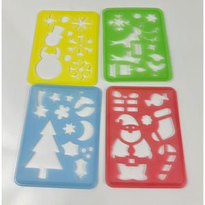 May include: Four colorful plastic stencils with Christmas-themed cutouts. The stencils are yellow, green, blue, and red. Designs include a snowman, snowflakes, a Christmas tree, Santa Claus, and candy canes.