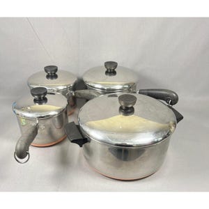 May include: A set of four stainless steel cooking pots with copper-coloured bases and matching lids. Each pot features a black handle and a black knob on the lid. The collection includes various sizes, all with a polished metallic finish and dark accents.