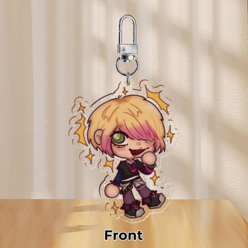 May include: A clear acrylic charm depicting a cartoon character with blonde hair, pink highlights, and green eyes. The character is winking and wearing a black top with purple trousers. The charm has a silver keychain and the word "Front" is printed at the bottom.