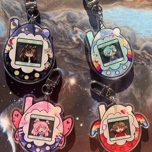 May include: Four acrylic keychains shaped like digital pet devices, each featuring a unique character illustration on the screen. The keychains include a dark purple, a light blue, a pink, and a red design, all equipped with silver metal clasps.