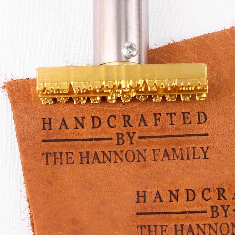 May include: A gold-colored metal branding iron with a cylindrical handle and a rectangular head featuring raised letters. The tool is pressed into a piece of tan leather, leaving a clear, dark imprint that reads: HANDCRAFTED BY THE HANNON FAMILY.