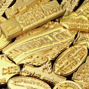 May include: A collection of gold-colored metal embossing stamps featuring various raised text and designs. Visible text includes CHURCH FOUNDATION, AND LONGENDYKE, and SALOON. The stamps have polished surfaces with intricate lettering and patterns.
