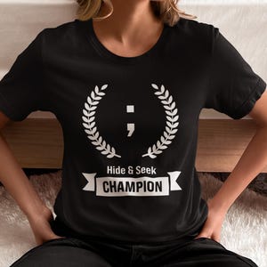 May include: Black t-shirt with a white graphic of a semicolon inside a laurel wreath. The text "Hide & Seek CHAMPION" is below the wreath.