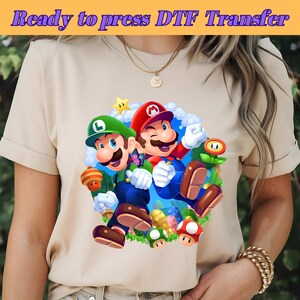 Pode incluir: Uma camiseta bege com um gráfico colorido de Mario e Luigi do videogame Super Mario Bros. A imagem inclui o texto "Ready to press DTF Transfer" no topo. O design mostra os personagens com vários elementos do jogo.