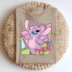 May include: A beige t-shirt with a cartoon image of Stitch, a blue alien character from the Disney movie Lilo & Stitch, with pink, blue, and green accents. The text "Angel" is written in pink letters.