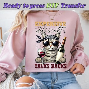 May include: Pink sweatshirt featuring a graphic of a cat wearing sunglasses and a bandana, holding a glass of wine and a bottle of wine. The text on the sweatshirt reads "Expensive, difficult, talks backs".