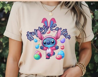 Puede incluir: Una camiseta beige con un diseño de orejas de conejo rosas y un personaje azul Stitch que lleva un disfraz de conejo rosa. El texto "Happy Easter" está escrito en una fuente colorida sobre el personaje. El personaje está rodeado de huevos de Pascua coloridos.