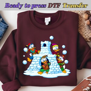 May include: Burgundy sweatshirt with a cartoon graphic of Donald Duck and his nephews playing in the snow. The image includes a snow fort and several snowballs. Text at the top reads "Ready to press DTF Transfer".