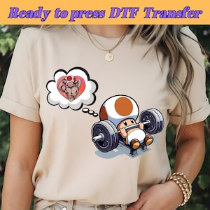 Op de afbeelding: Een beige t-shirt met een cartoon afbeelding van een paddenstoelfiguur die gewichten heft. Het personage droomt van een gespierde figuur in een hartvormige wolk.