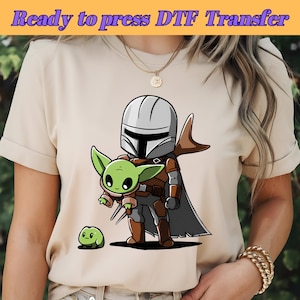 以下が含まれることがあります： ベージュのTシャツに、マンダロリアンキャラクターが小さな緑色のエイリアンを抱いている漫画の画像が描かれています。画像の上部には「Ready to press DTF Transfer」というテキストがあります。