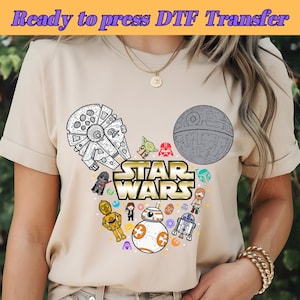 以下が含まれることがあります： ベージュのTシャツに、白とグレーのスターウォーズのテーマのデザインが施されています。デザインは、ミッキーマウスの頭のシルエットで、耳の部分にミレニアムファルコンとデススターが描かれています。"STAR WARS"の文字は金色の文字で書かれています。デザインには、ヨーダ、ダースベイダー、R2-D2、チューバッカなど、さまざまなスターウォーズのキャラクターも含まれています。