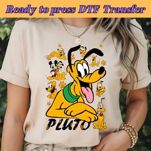 May include: A beige t-shirt with a graphic print of Pluto the dog from Disney. The graphic features Pluto in various poses with Mickey Mouse and other Disney characters. The text "PLUTO" is printed below the graphic.