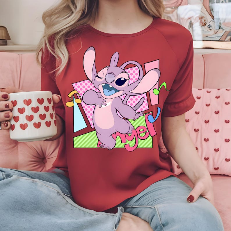 May include: Red t-shirt with a cartoon image of a purple Stitch character with blue eyes and a pink nose. The character is dancing and the text "Angel" is written in a colorful font.