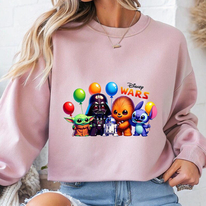 May include: Pale pink sweatshirt with a graphic of Star Wars and Disney characters, including Baby Yoda, Darth Vader, Chewbacca, and Stitch, holding balloons. The words "Disney Wars" are printed above the characters.