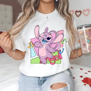 May include: White t-shirt with a cartoon image of Stitch from Lilo & Stitch. Stitch is purple and pink and is singing "Angel" in a colorful, geometric design.