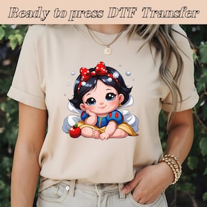 以下が含まれることがあります： ベビーのスノーホワイトがリンゴを持って座っている漫画の画像がプリントされたベージュのTシャツ。画像はTシャツの前面にプリントされています。