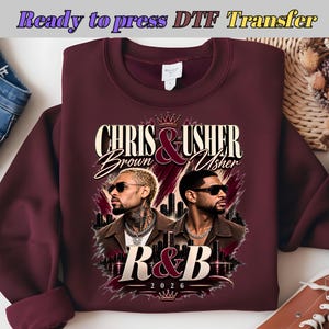 May include: A maroon crewneck sweatshirt featuring a graphic print of Chris Brown and Usher wearing sunglasses. The text reads "CHRIS & USHER", "Brown", "Usher", "R&B", and "2026". The design includes a crown icon, a city skyline silhouette, and brushstroke accents.