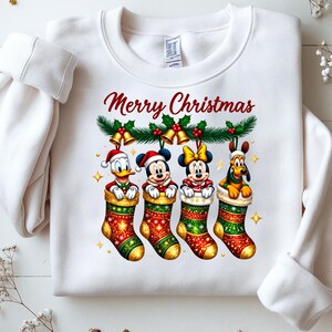 May include: White sweatshirt with a festive "Merry Christmas" design. Features Donald Duck, Mickey Mouse, Minnie Mouse, and Pluto in Christmas stockings. The design includes holly, bells, and gold stars.